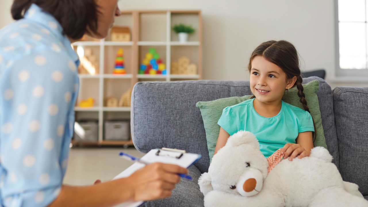 young girl sitting on couch holding stuffed bear and talking with caring therapist